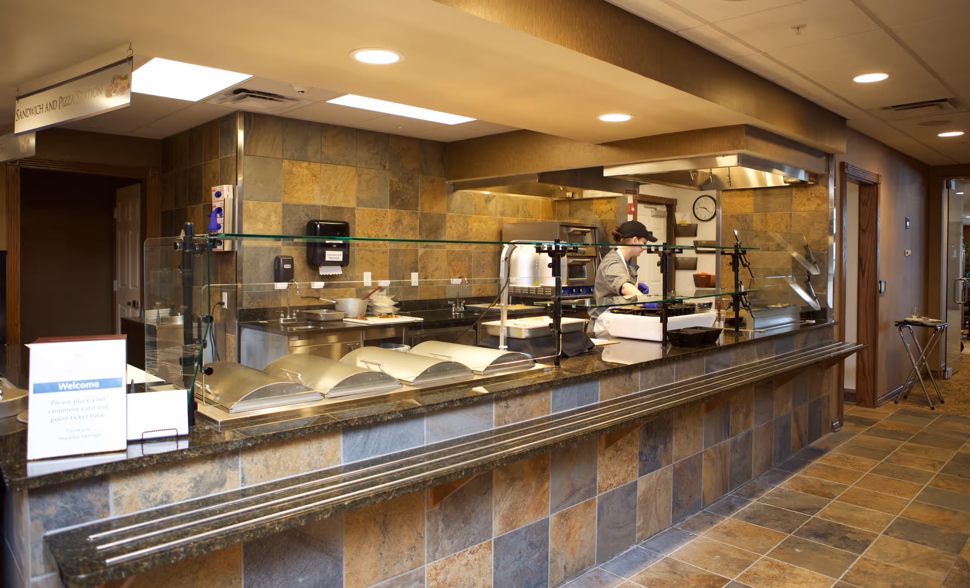 A serving counter area in a kitchen with a person working behind a glass partition. The counter has several covered food trays and a sign instructing guests to place their tickets in a box. The walls and floor are tiled with brown and beige stone tiles, and there is a clock on the wall in the background.