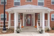 Front entrance of a red brick building with a white columned portico, double doors labeled 2000, and potted white flowers and pumpkins.