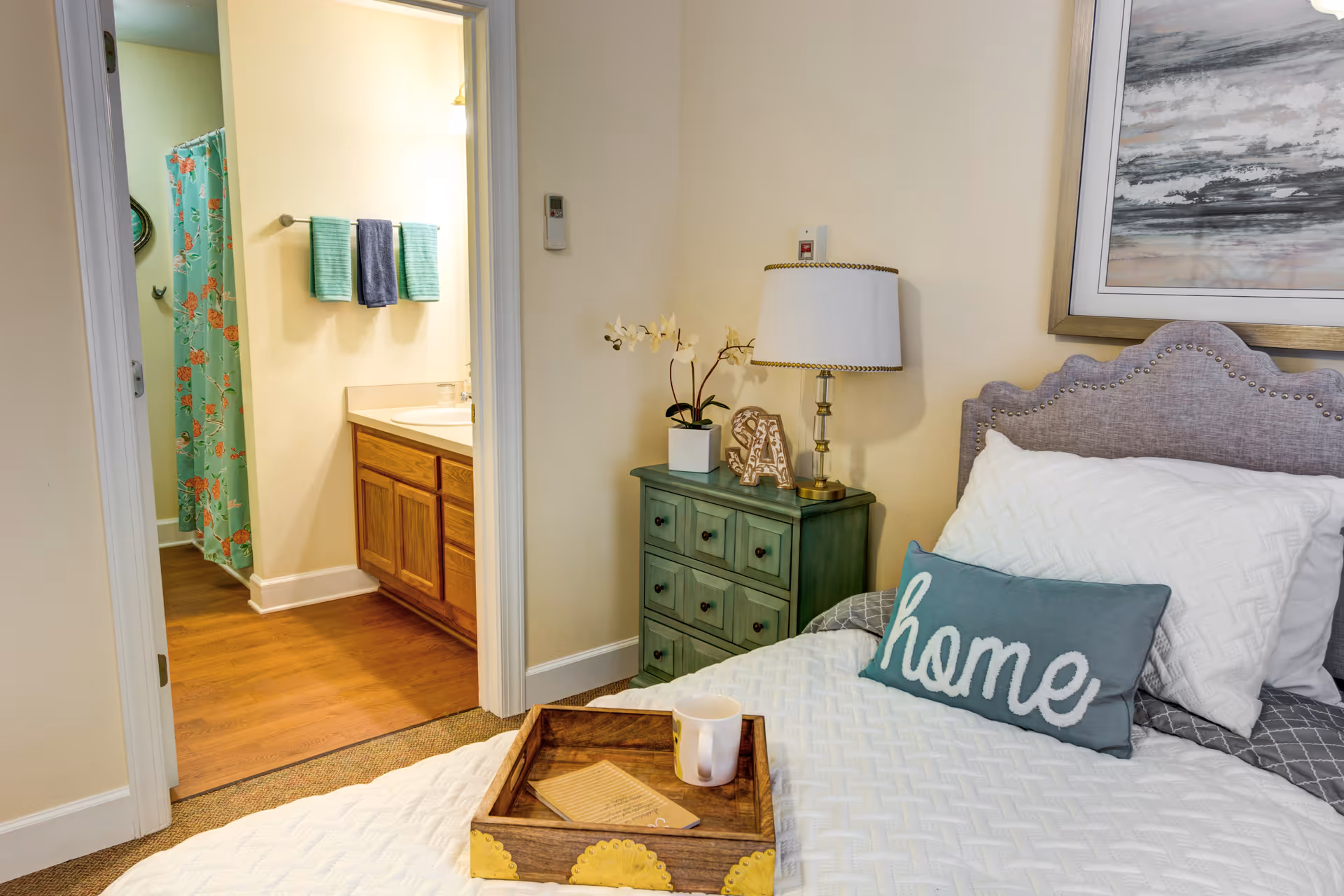 A bedroom with a made bed featuring a 'home' pillow, green nightstand with a lamp, and an open doorway showing an adjacent bathroom.