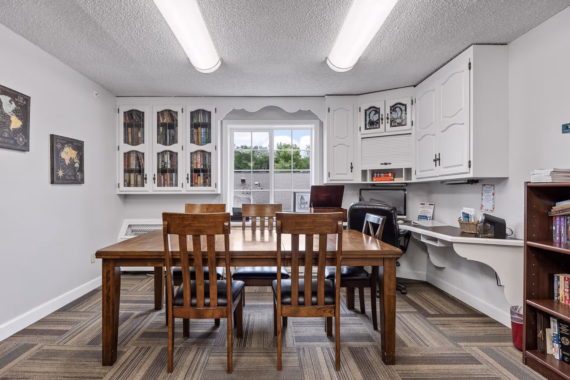 A room with a wooden table and six wooden chairs with black cushions. Behind the table is a window with a view of trees and rooftops. On the left wall, there are two framed maps, and above the window are white cabinets with glass doors containing books. On the right side, there is a built-in desk area with a black office chair, computer monitor, and various office supplies. A bookshelf with books is visible on the far right.