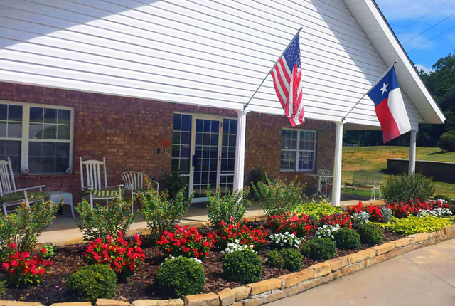 Front entrance porch with rocking chairs, landscaped flowerbeds, and American and Texas flags.