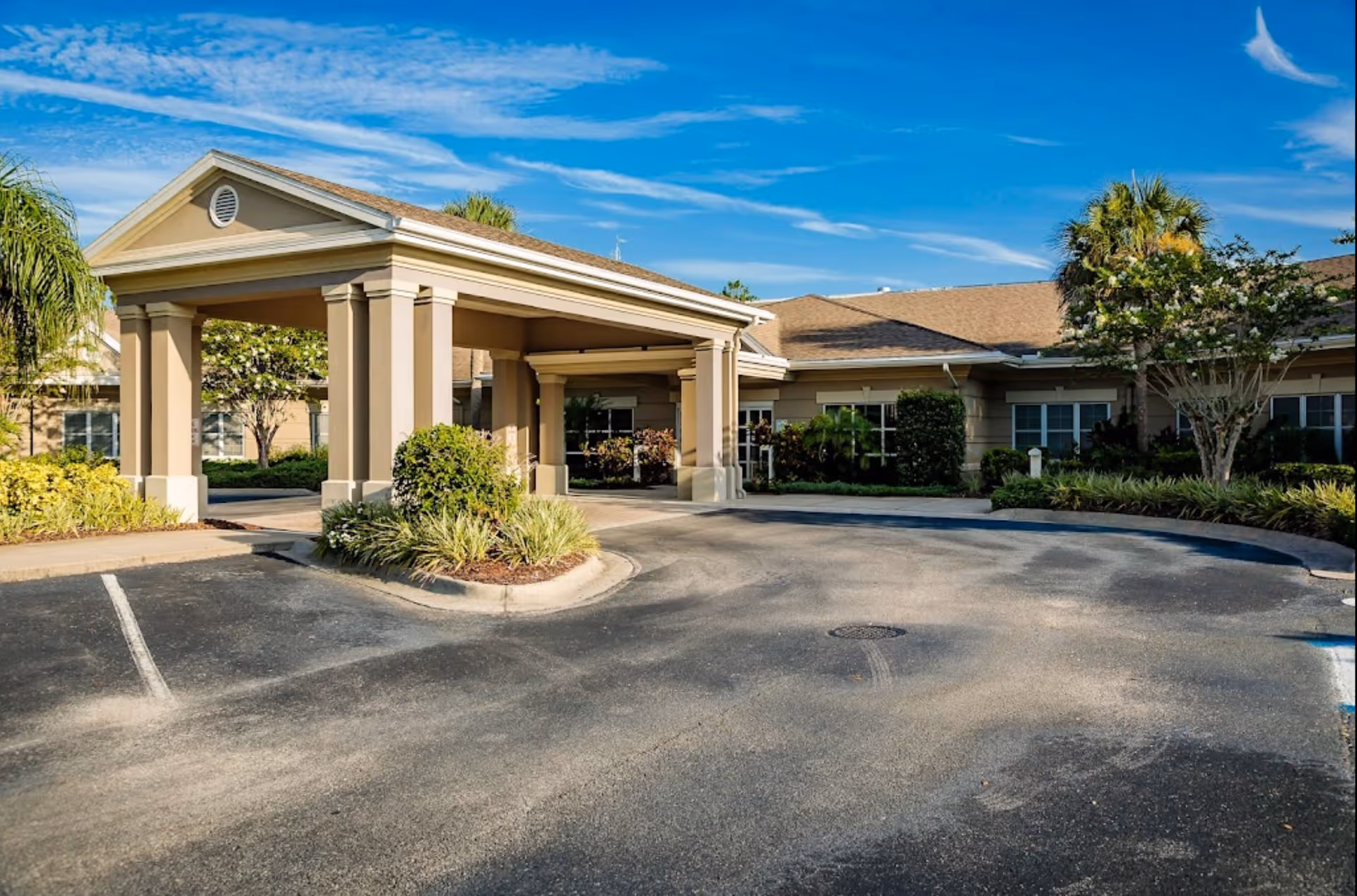 The front entrance of a one-story senior care building with a covered porte-cochère, driveway, and landscaped shrubs under a blue sky.