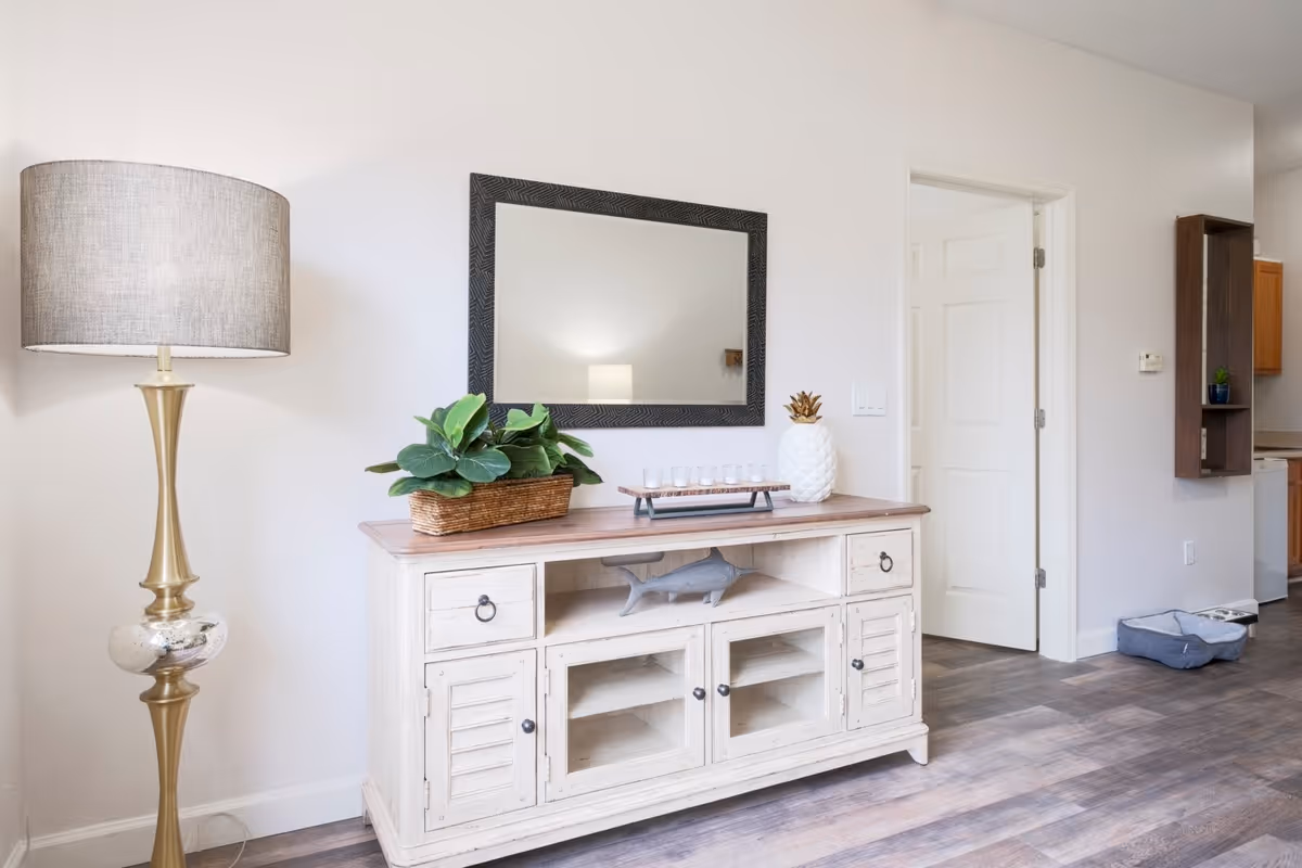 A bright living-room area featuring a white distressed console table with decor and mirror, a tall floor lamp, and an open doorway to the kitchen.