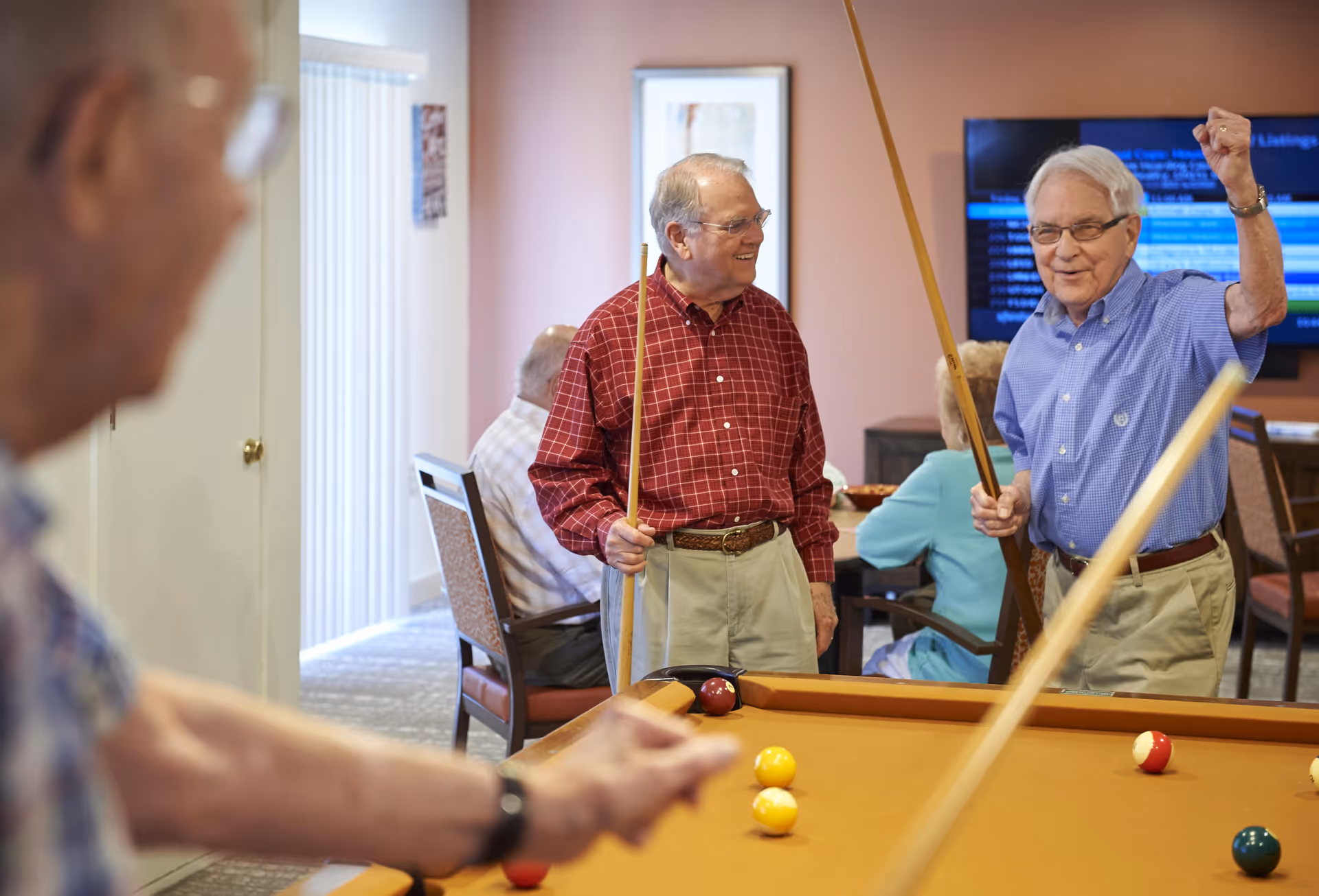 Three elderly men playing pool in a common area of a senior living facility. Two men are standing near the pool table holding cues, one smiling and the other raising his fist in excitement. Another man is in the foreground aiming a shot. In the background, two elderly people are seated and a television screen is mounted on the wall.