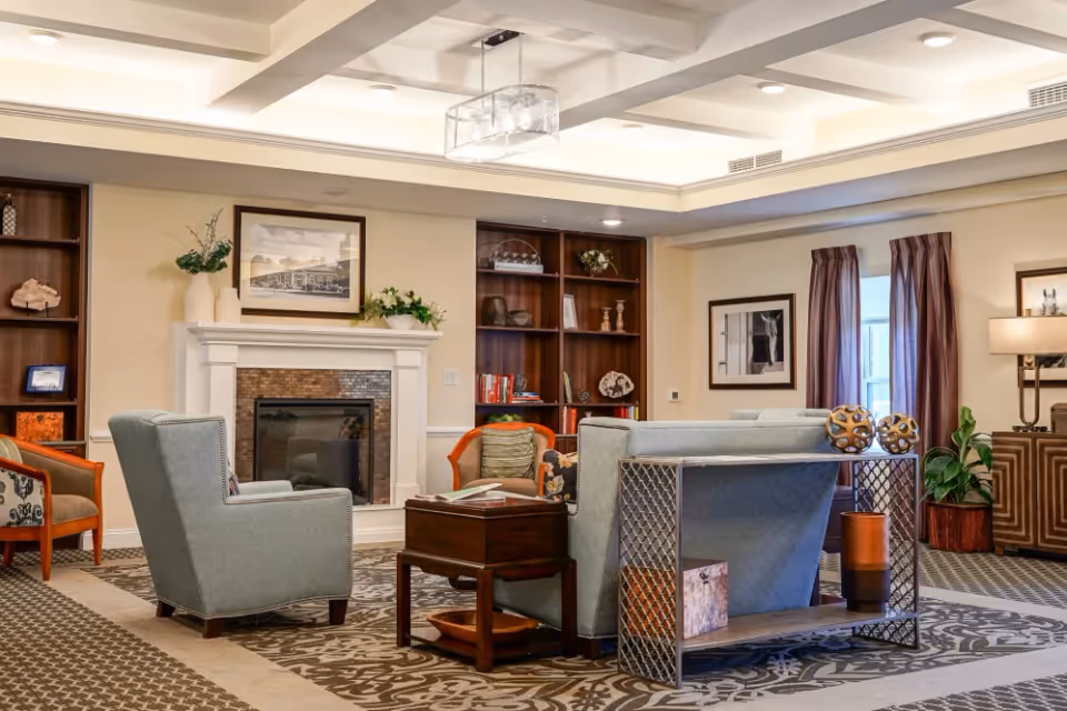 A cozy lobby area featuring comfortable seating, a fireplace, and decorative bookshelves with various items. The room is well-lit with natural light coming through the windows, and there are plants and artwork enhancing the ambiance.