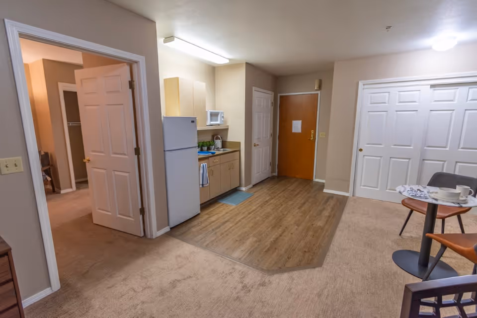 Interior view of a senior living apartment showing a small kitchenette with a refrigerator, microwave, sink, and cabinets. The floor transitions from wood in the kitchen area to carpet in the living space. There is a small round table with two chairs set with cups and plates. Multiple doors are visible, including a wooden entrance door and white closet doors.
