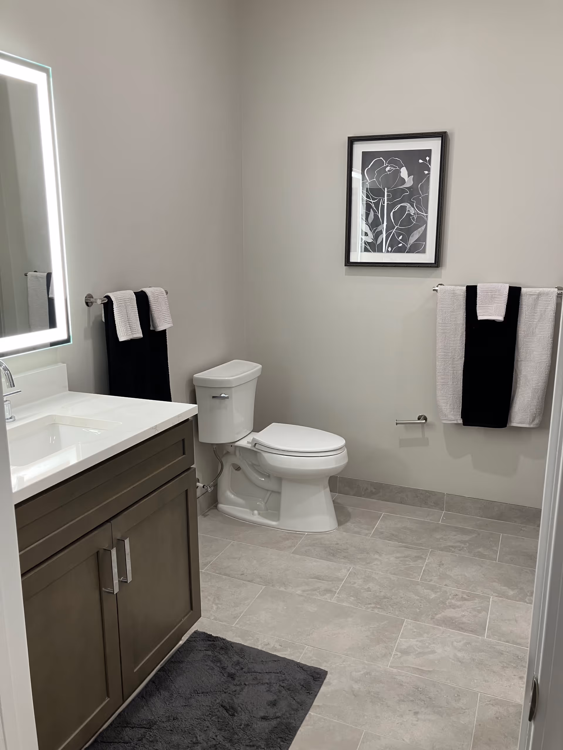 A clean and modern bathroom with a white toilet, a vanity with a white countertop and a lit mirror above it. There are black and white towels hanging on towel racks on the walls, a framed black and white floral artwork, and a dark gray bath mat on the tiled floor.