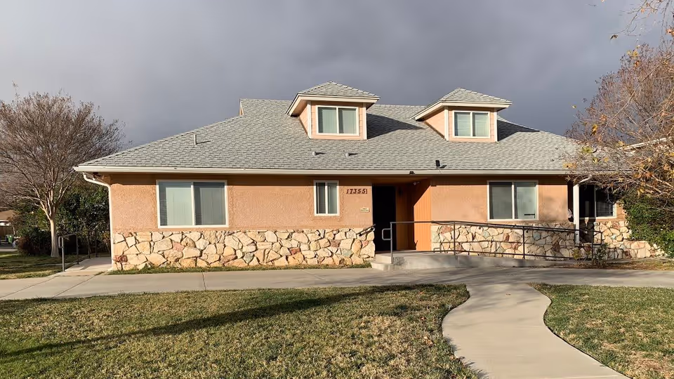 Single-story building with a gray shingled roof, beige walls with stone accents along the bottom, and two dormer windows. There is a concrete walkway leading to the entrance with a handrail ramp. Leafless trees and grass surround the building under a cloudy sky.