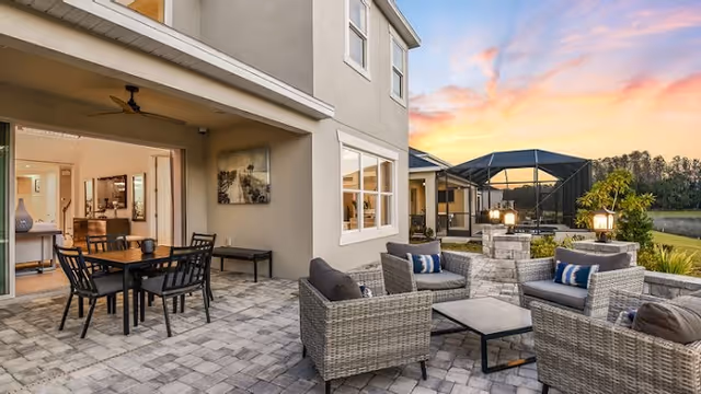 Outdoor patio area of a residential facility at sunset featuring a seating arrangement with wicker chairs and cushions around a low table, a dining table with chairs under a covered section, and a screened pool enclosure in the background near a body of water.