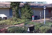 Single-story facility front with a walkway, garden shrubs, a birdbath, and potted plants.