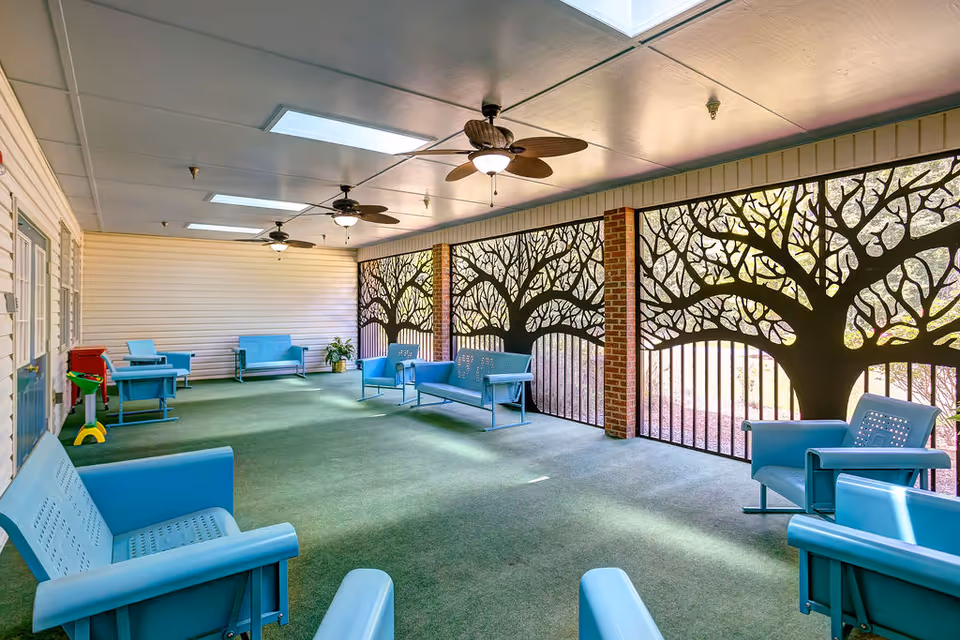 A covered outdoor seating area with light blue metal benches and chairs arranged on a green carpet. The space features ceiling fans with lights and decorative metal panels with tree designs along one side, allowing natural light to filter through.