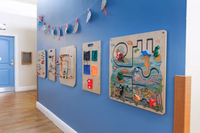 Hallway with a blue wall lined with several mounted wooden activity panels and a decorative garland, with a blue door visible at the end.