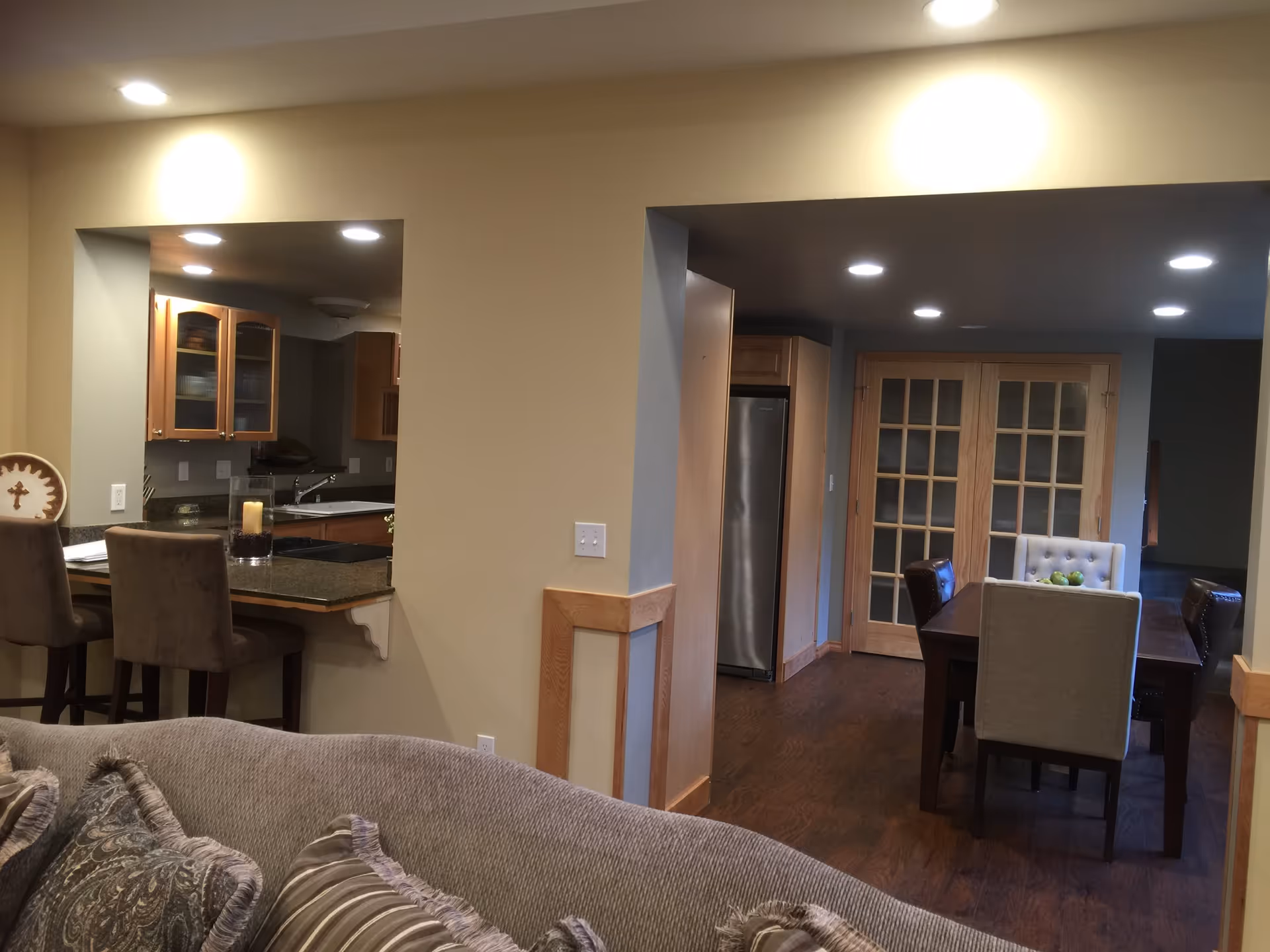 Interior view of a senior living facility showing a cozy living room with patterned cushions on a sofa in the foreground, a kitchen area with wooden cabinets and a countertop with bar stools on the left, and a dining area with a wooden table and chairs in the background. The dining area has glass-paneled double doors and a stainless steel refrigerator is visible.