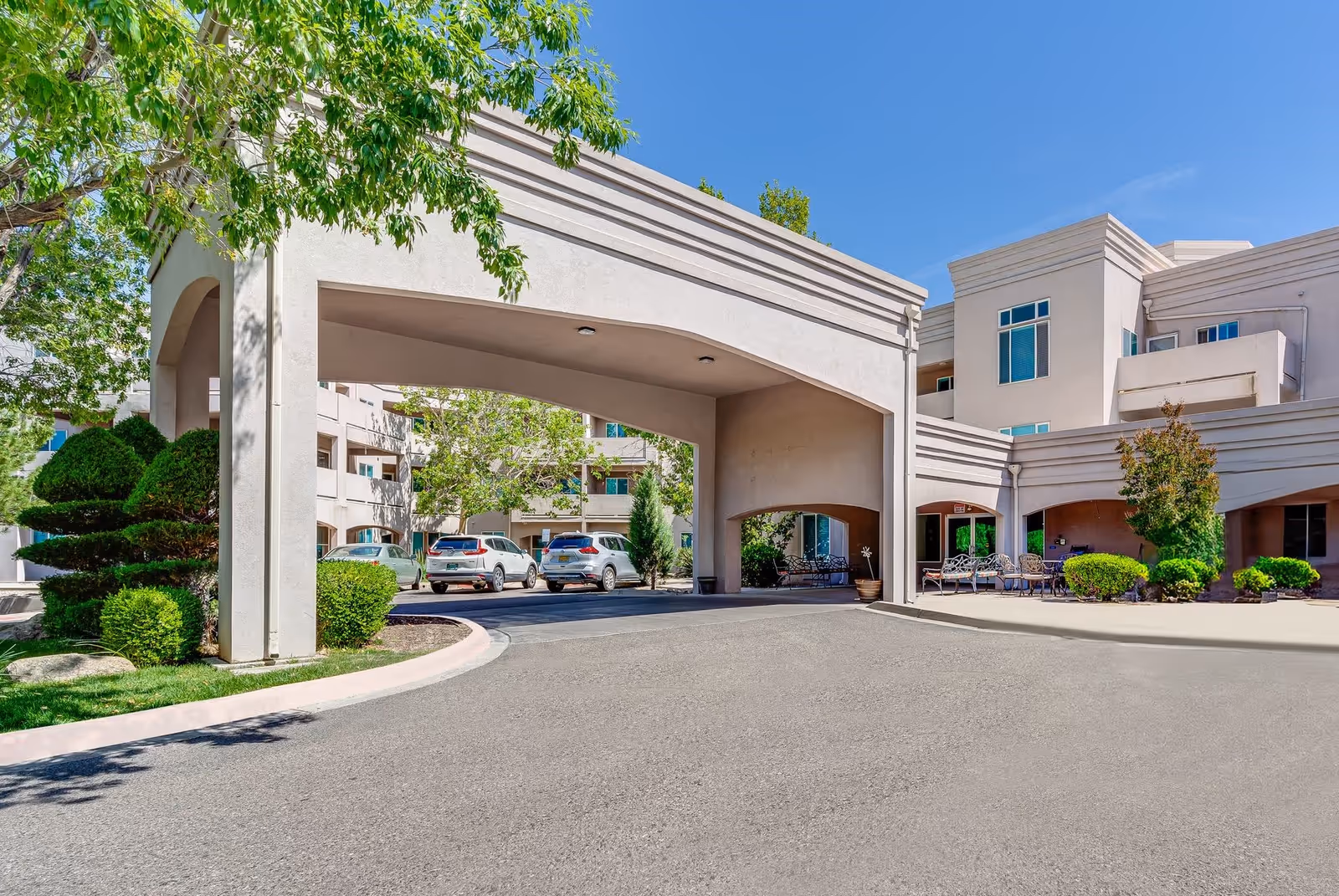 Front entrance and porte-cochère of a light-colored multi-story senior living building with parked cars and landscaped greenery under a clear blue sky.