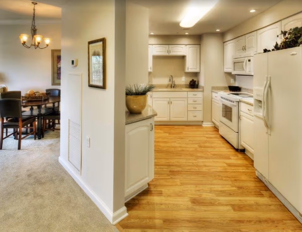 Interior view showing a kitchen with white cabinets, a refrigerator, stove, microwave, and wooden flooring. Adjacent to the kitchen is a dining area with a wooden table and chairs, carpeted floor, and a chandelier overhead.