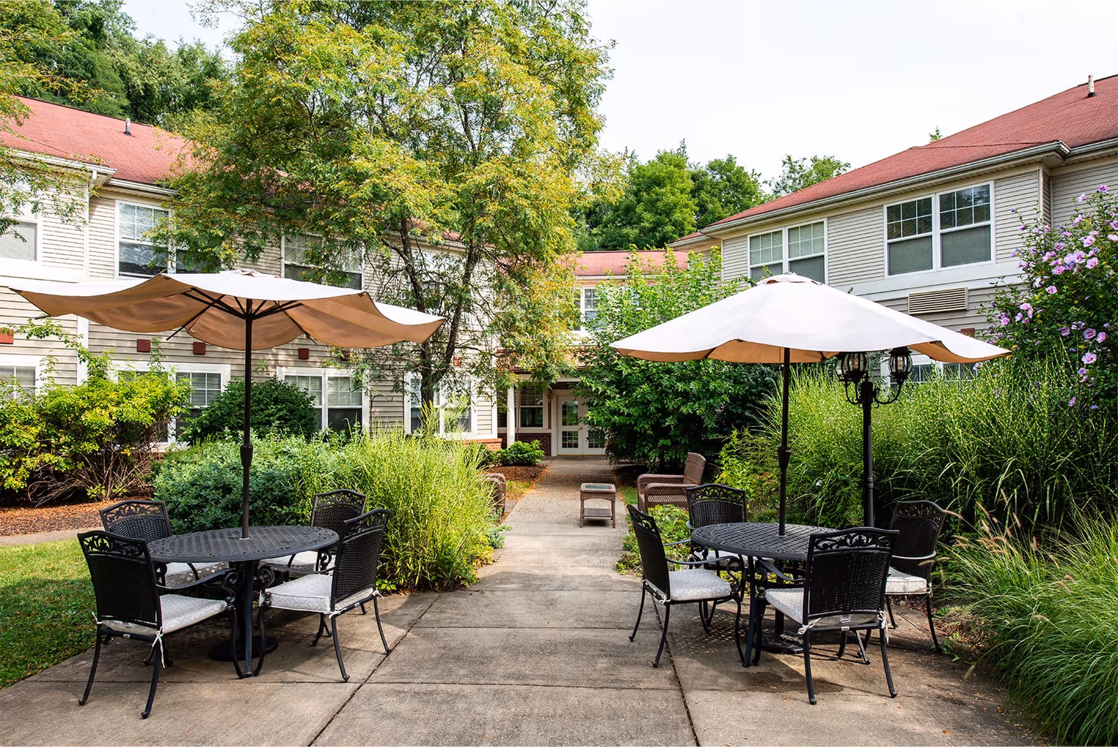 Outdoor patio area at Hawthorne Woods by New Perspective featuring two round black metal tables with umbrellas and cushioned chairs, surrounded by lush greenery and flowering bushes, with a two-story building with red roofs in the background.