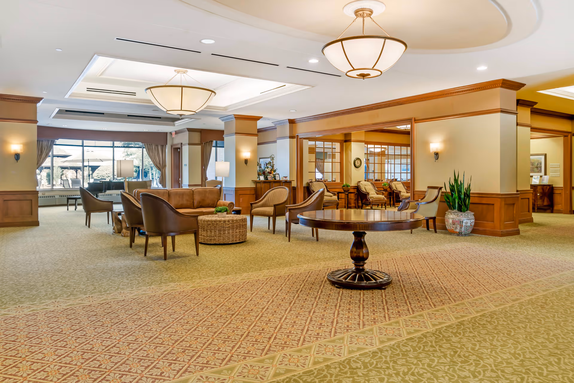A spacious and well-lit senior living facility common area with comfortable seating including sofas and armchairs arranged around a round wicker coffee table. The room features large windows with curtains, warm beige walls with wooden paneling, decorative ceiling lights, and a round wooden table in the foreground. There are plants and framed pictures adding to the cozy atmosphere.