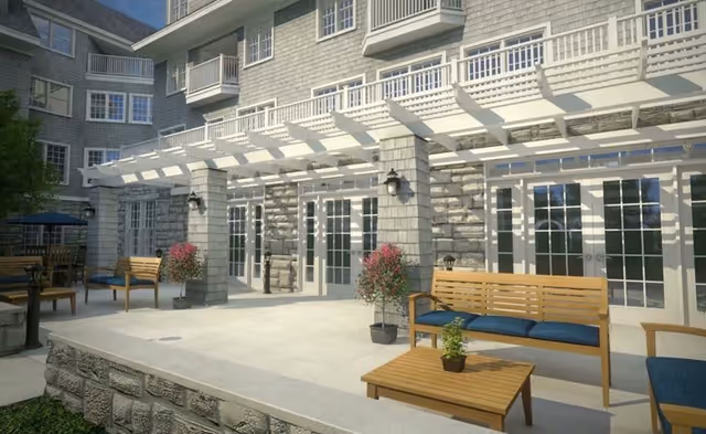 Outdoor patio area of a senior living facility with wooden benches and chairs featuring blue cushions, a wooden coffee table with a small potted plant, stone pillars supporting a white pergola, and large glass doors leading inside the building.