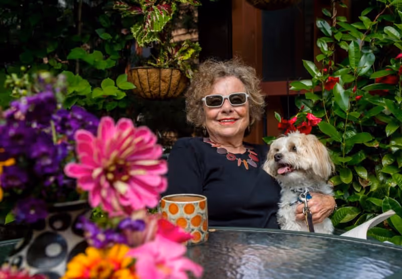 An elderly woman wearing sunglasses and a black top sits outdoors at a glass table surrounded by vibrant flowers and greenery. She is smiling and holding a small white dog with light brown patches. A colorful mug is on the table in front of her.