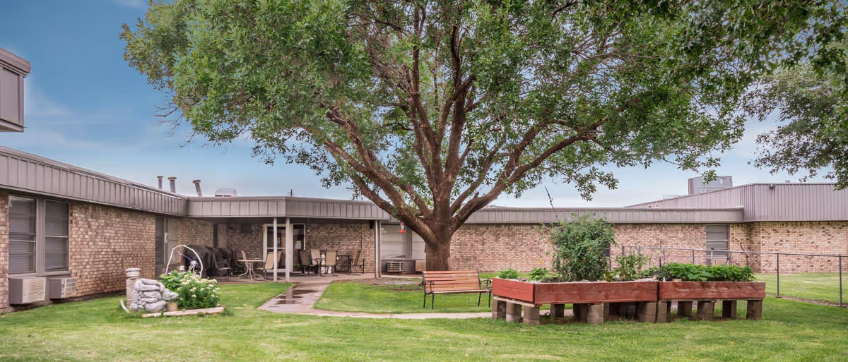Outdoor courtyard area of Prairie Acres Nursing Home/Friona Heritage Estates featuring a large tree in the center, a wooden bench, raised garden beds, a small water fountain, and a covered patio with tables and chairs. The building is a single-story brick structure surrounding the courtyard.