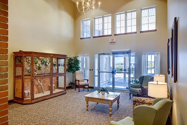 Sunlit senior living facility lobby with armchairs, a coffee table, a chandelier, tall windows above the entrance, and a wooden display cabinet against the wall.