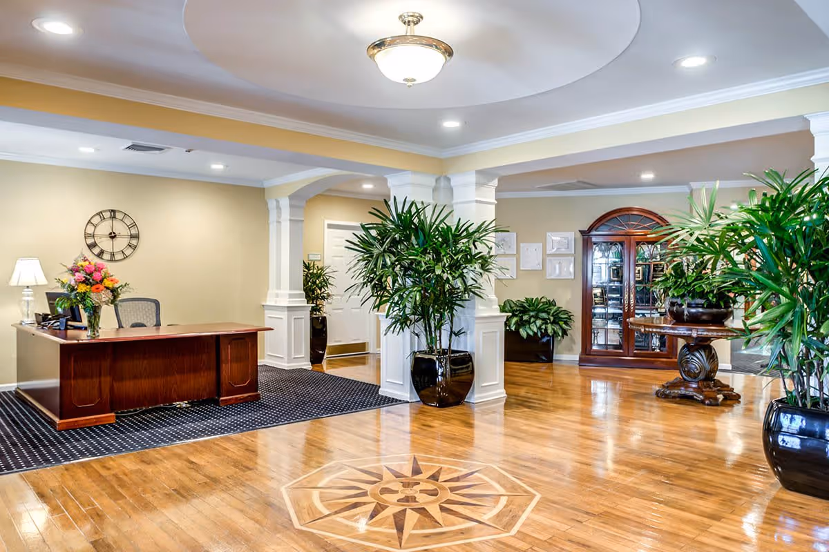 A spacious and well-lit reception area with a wooden desk adorned with a bouquet of flowers and a lamp. The floor features polished wood with a decorative compass design in the center. There are several large potted plants, white columns, and a wooden cabinet with glass doors in the background. The walls are painted a soft beige color, and a round clock hangs above the desk.
