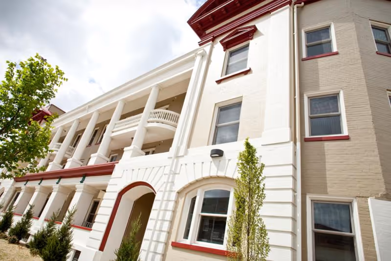Exterior view of a multi-story building with white and beige brick walls, red trim around windows and roofline, and white columns supporting balconies. Small trees and shrubs are planted along the building's base under a partly cloudy sky.