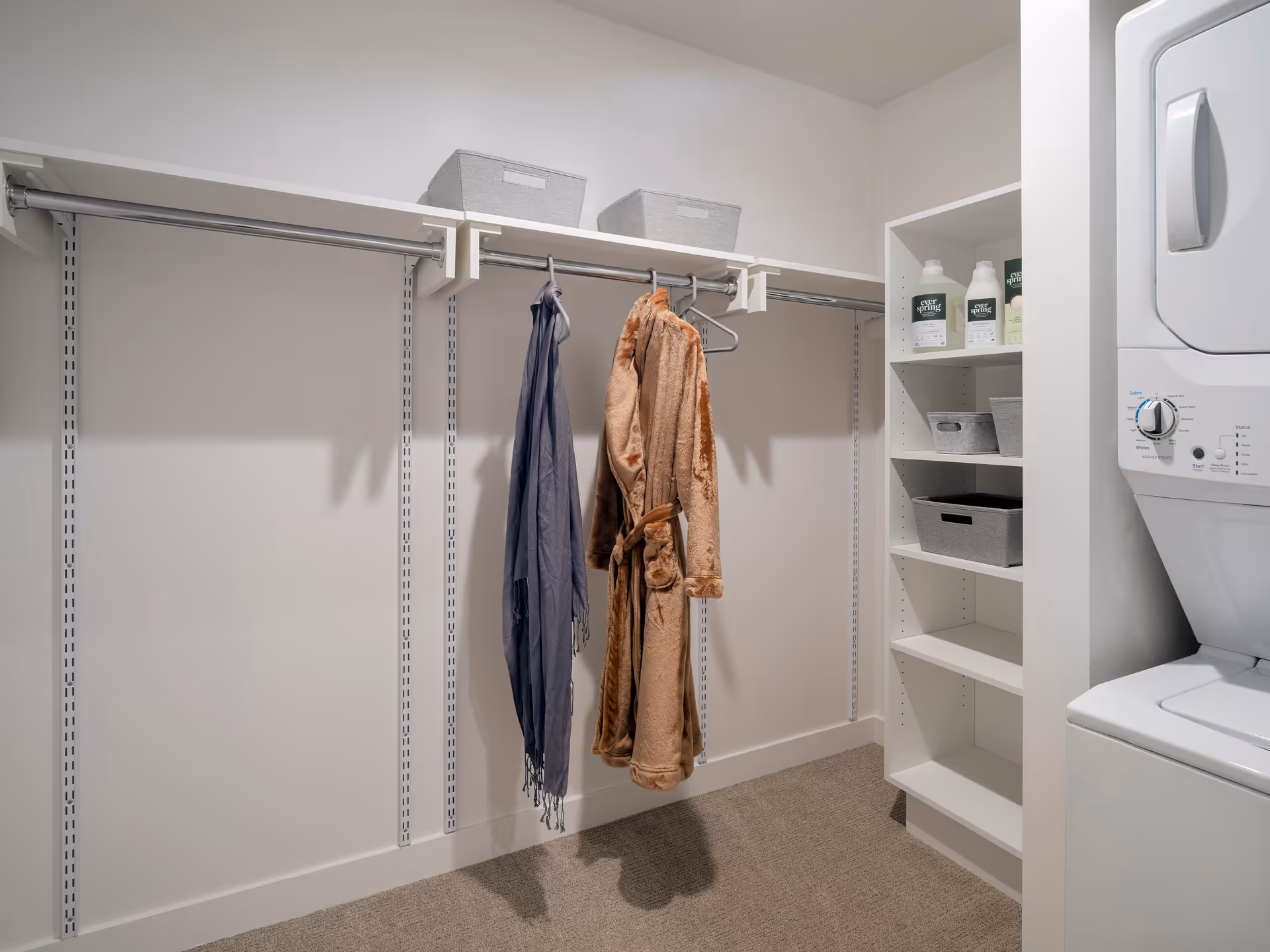 A small laundry room with a stacked washer and dryer on the right, white shelving with laundry supplies, and a closet rod with two hanging garments and storage bins on the top shelf.