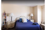A cozy bedroom with a blue-covered bed flanked by matching nightstands and lamps, a bookshelf, and a chair by the window.