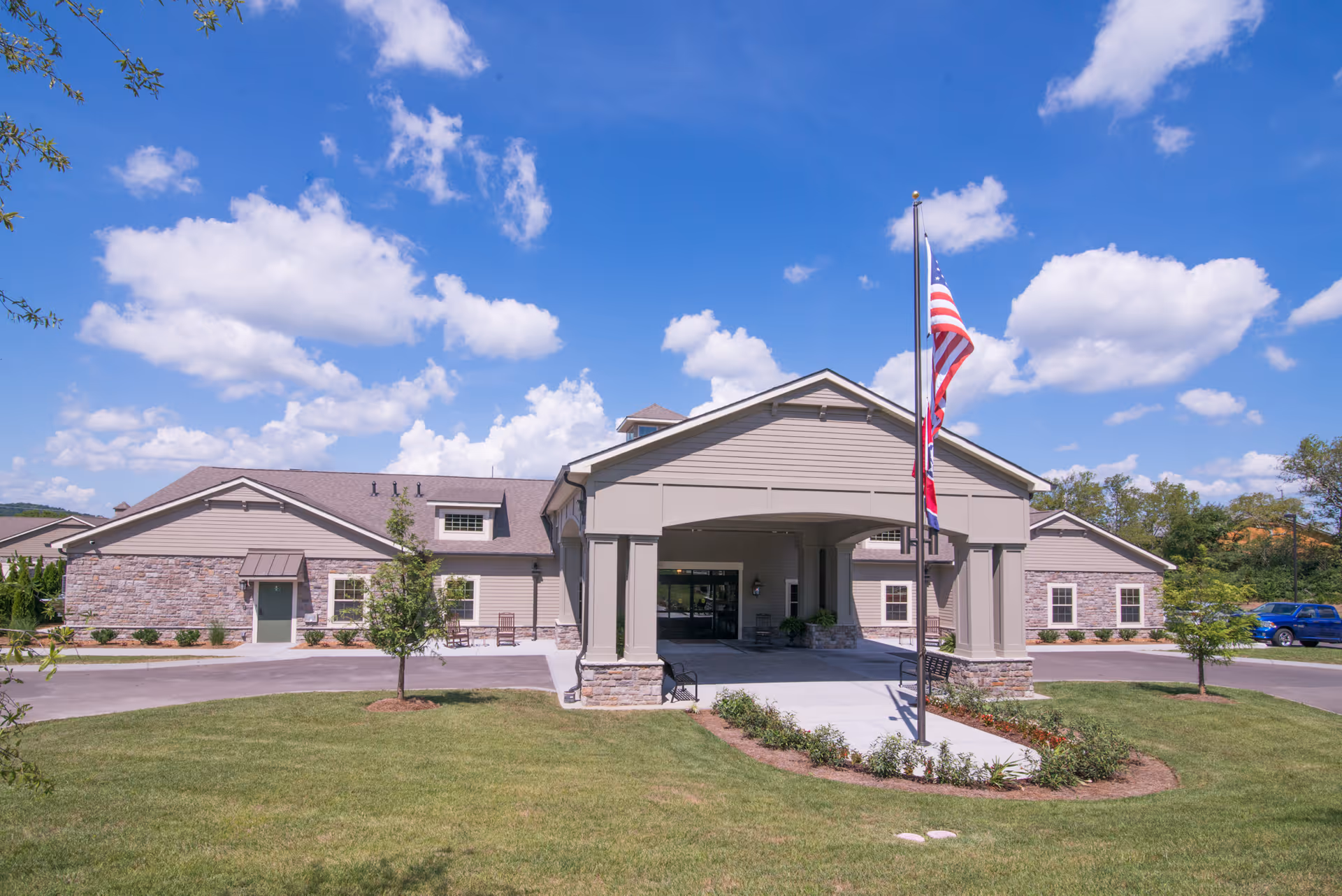 Front exterior view of a single-story senior living facility building with a covered entrance, an American flag on a flagpole, a well-maintained lawn, and a bright blue sky with scattered clouds.