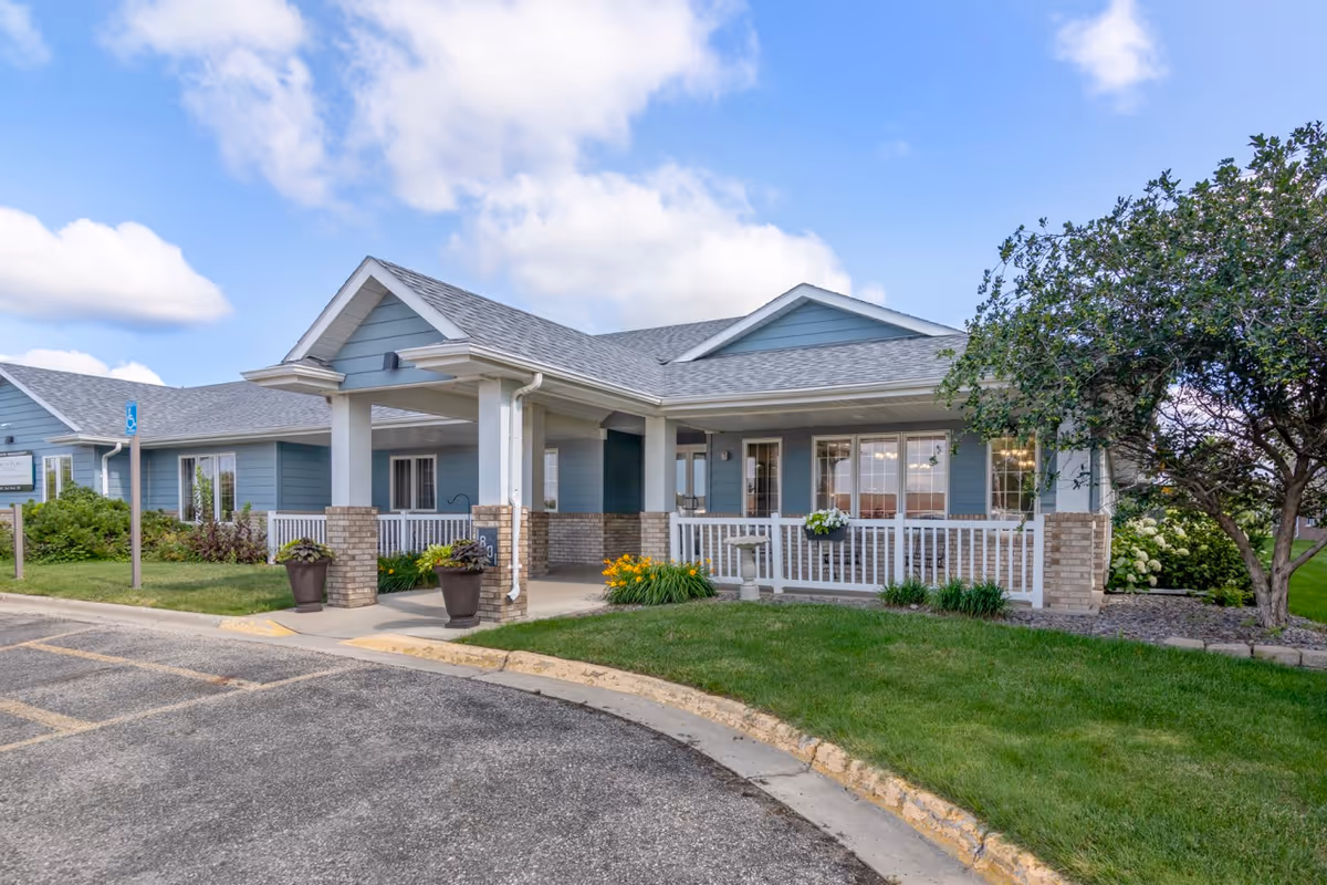 Covered front entrance and porch of a light-blue single-story senior living building surrounded by a manicured lawn and potted plants.