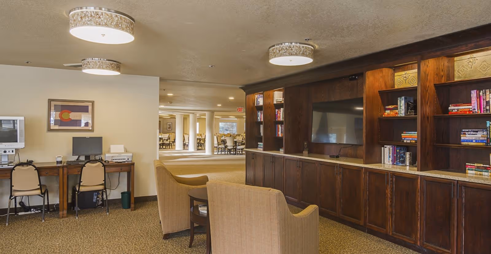 Interior view of a senior living facility common area with two beige armchairs facing a large wooden entertainment center with a flat-screen TV and bookshelves. To the left, there is a desk with two chairs, a computer, and a printer. In the background, a dining area with tables and chairs is visible.