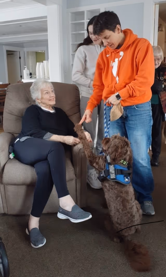 An elderly woman in a recliner shakes a brown service dog's paw while a person in an orange hoodie holds the leash in a communal living room.