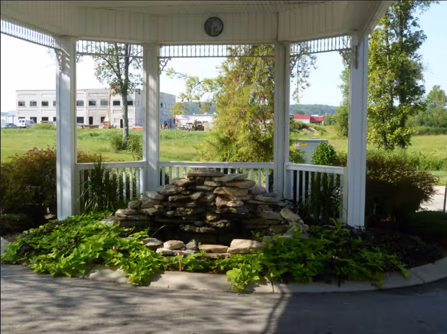 A white gazebo sheltering a stone water feature surrounded by green plants with a grassy field and buildings in the background.