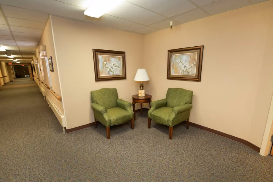A corner seating area in a senior living facility hallway with two green upholstered armchairs, a small round wooden table with a lamp and decorative items, beige walls, two framed floral paintings, and a long carpeted corridor with handrails and windows.