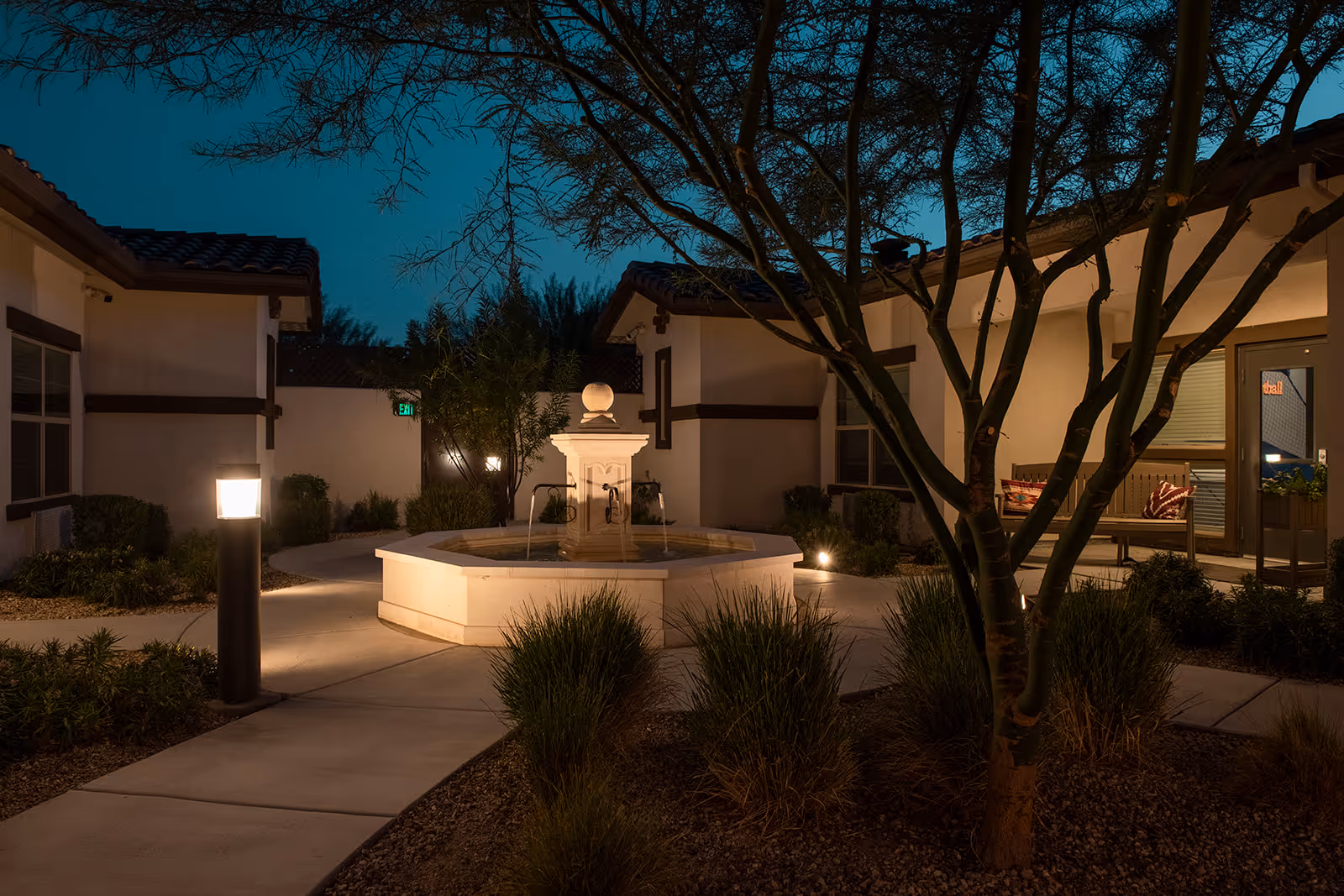 Nighttime view of an outdoor courtyard area at a senior living facility with a central water fountain surrounded by pathways, landscaping, and illuminated lamp posts. The courtyard is bordered by single-story buildings with tiled roofs and windows, and there is a bench with cushions near one of the building entrances.
