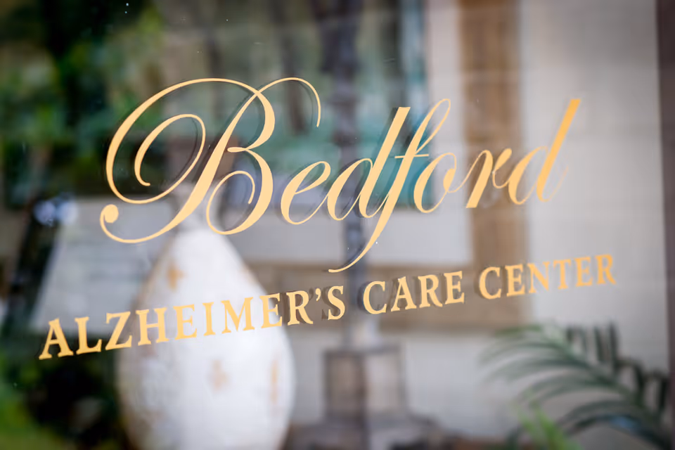 Close-up view of a glass door or window with gold lettering that reads 'Bedford Alzheimer's Care Center', with blurred indoor plants and decor visible in the background.