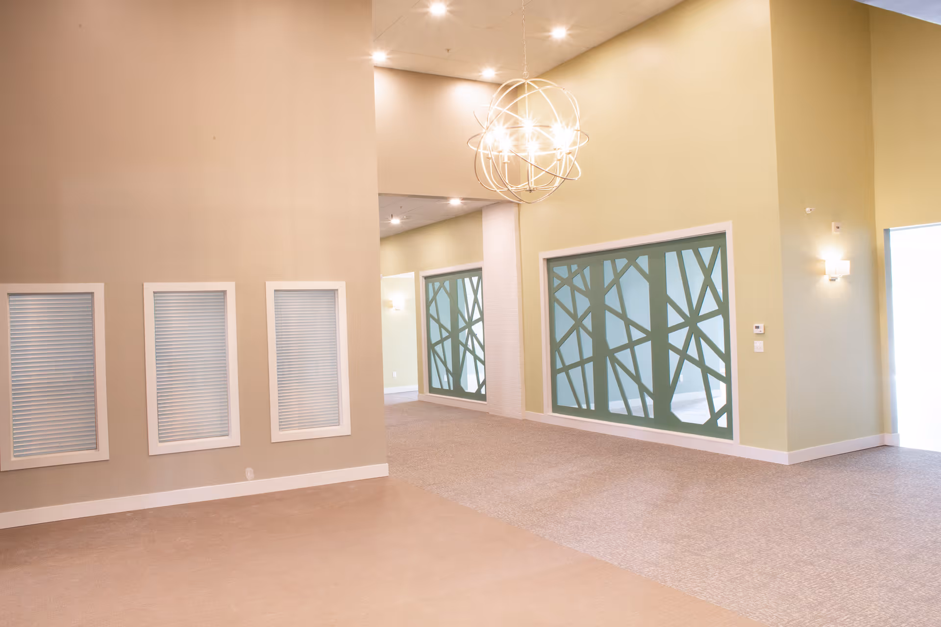 Interior view of a senior living facility hallway with beige and light green walls, three small windows with closed blinds on the left wall, decorative large windows with abstract tree branch patterns on the right wall, a modern spherical chandelier hanging from the ceiling, and carpeted floors.