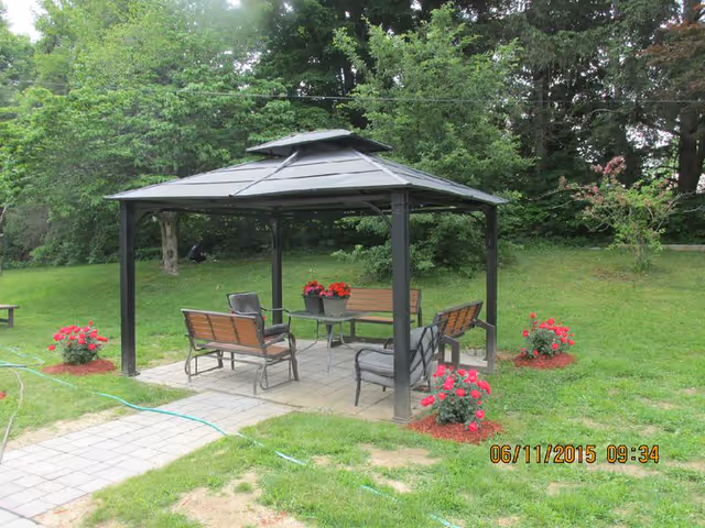 Outdoor garden area with a black metal gazebo featuring a peaked roof. Under the gazebo are two wooden benches, two chairs, and a small round table with a flower pot containing red flowers. The gazebo is surrounded by green grass, trees, and additional flower beds with red flowers. A paved walkway leads to the gazebo.