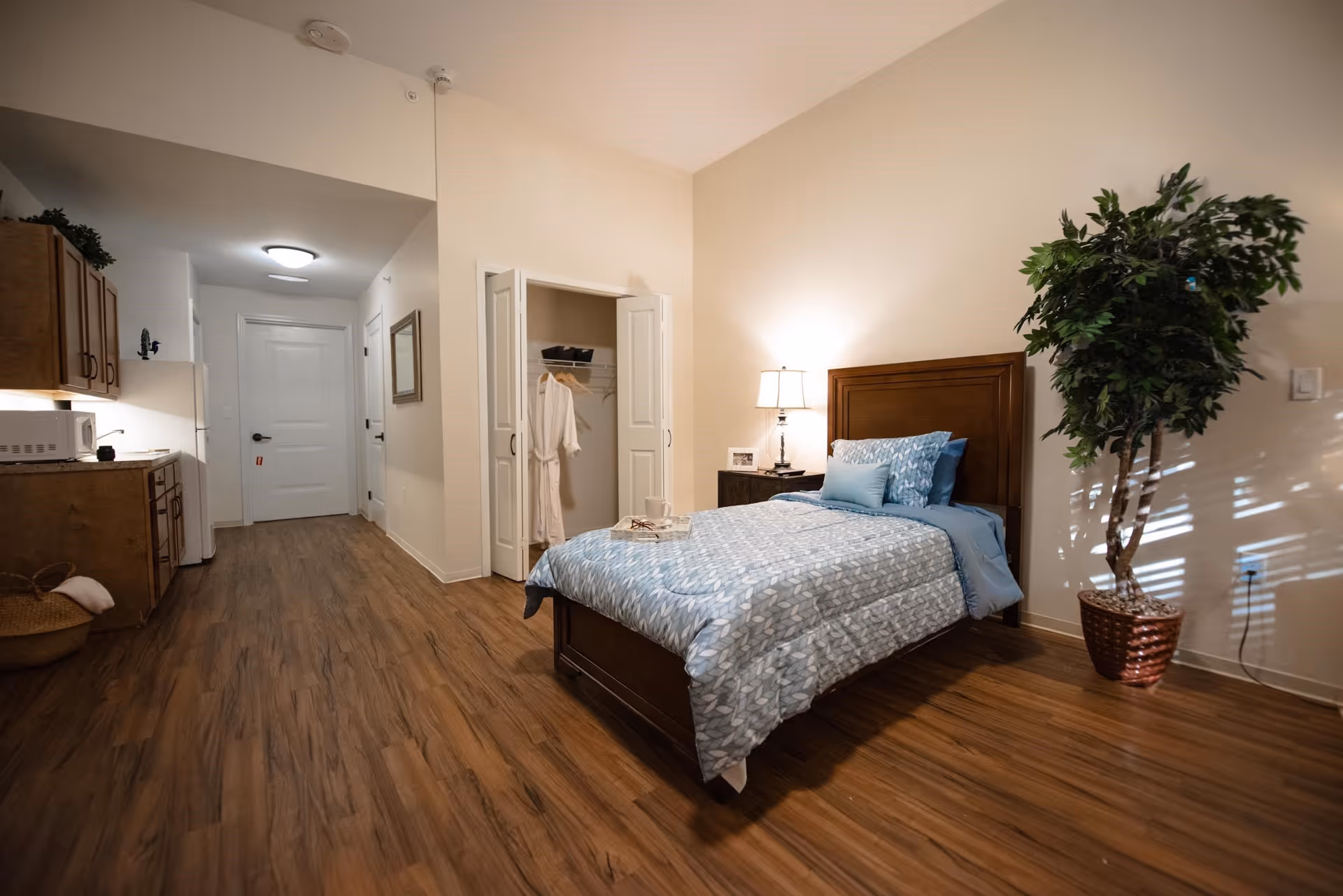 A senior living facility room featuring a single bed with blue patterned bedding and pillows, a wooden nightstand with a lamp, an open closet with hanging robes, a large potted plant, and a kitchenette area with cabinets, a microwave, and a refrigerator. The room has wood flooring and neutral-colored walls.