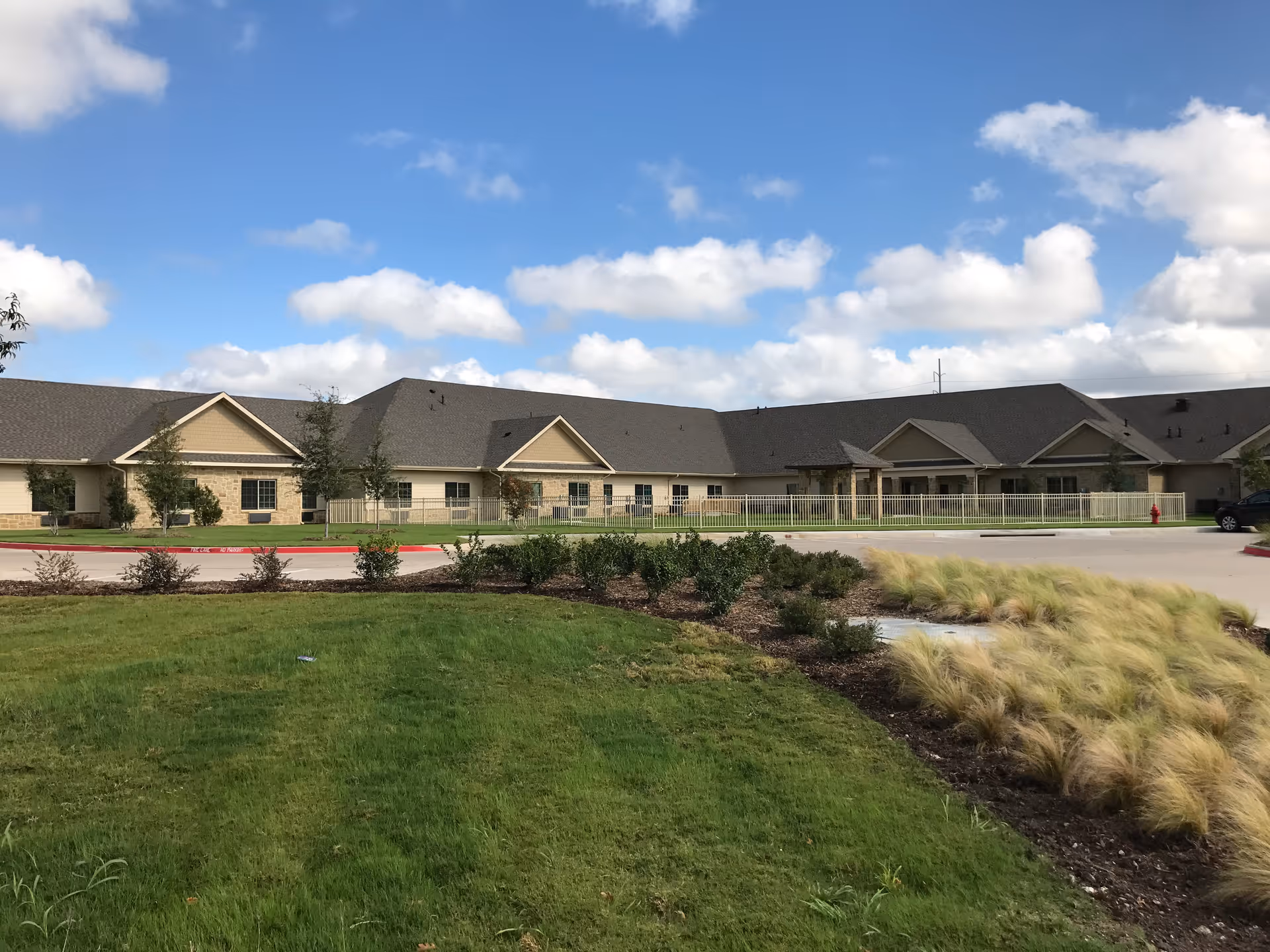 Exterior view of a single-story healthcare and rehabilitation facility building with beige walls and a dark gray roof under a partly cloudy blue sky. The foreground features a well-maintained lawn, landscaped bushes, and ornamental grasses along a curved driveway.
