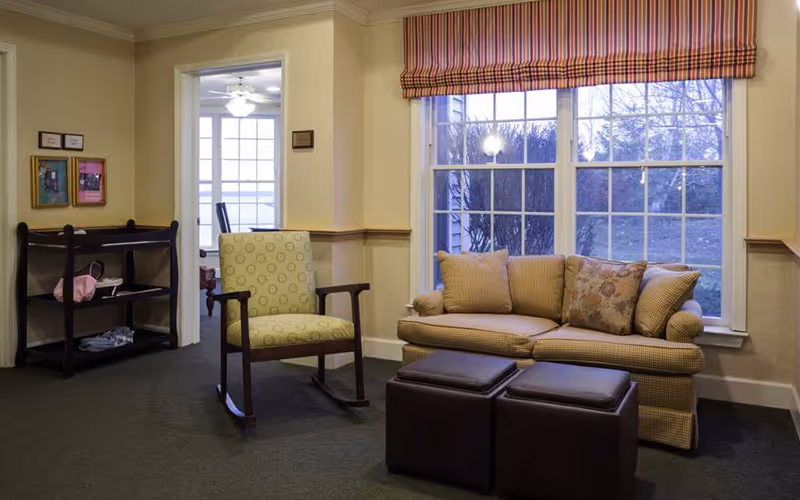 A cozy living room area with a patterned rocking chair, a beige sofa with multiple cushions, and two dark brown ottomans in front of the sofa. The room has large windows with striped valances, beige walls with a chair rail, and a small shelving unit with framed pictures on the wall.