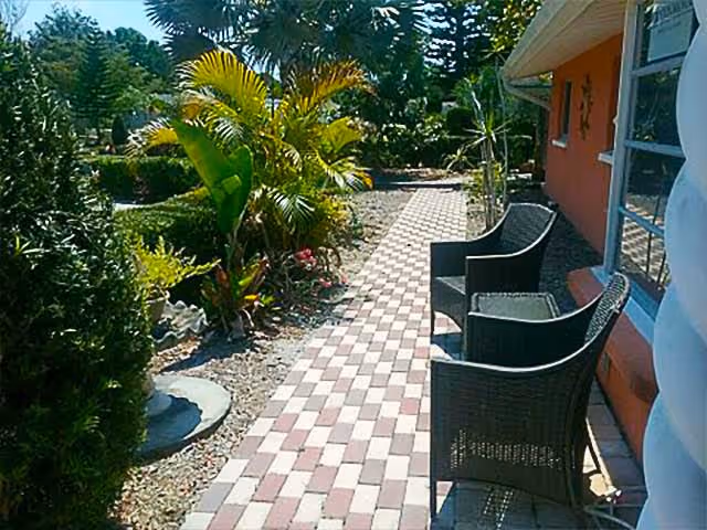Outdoor patio area with two black wicker chairs placed on a checkered brick pathway next to a building with orange walls and windows. The pathway is bordered by various green plants and trees under a sunny sky.