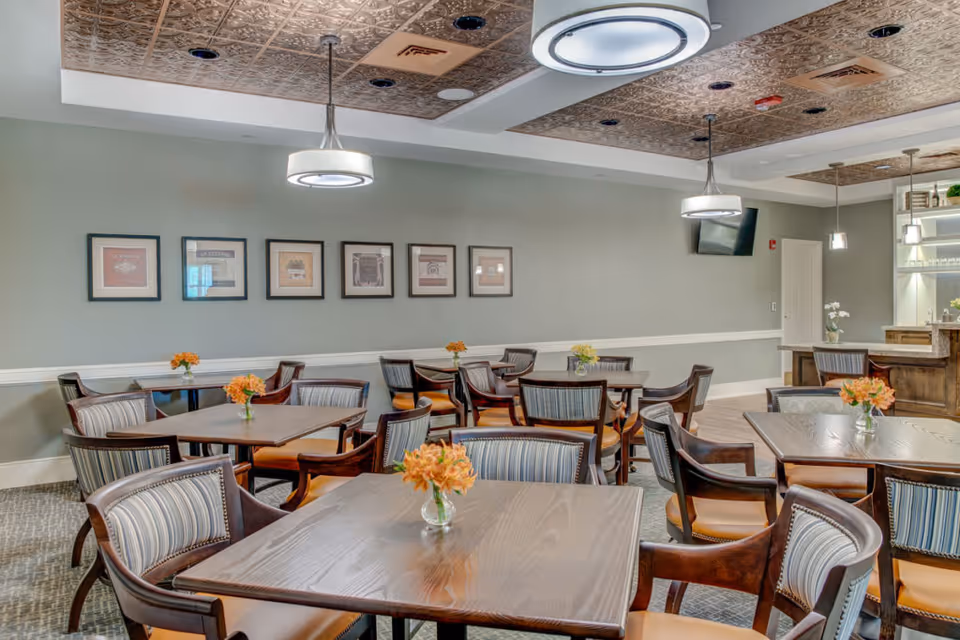 Dining room with multiple wooden tables and upholstered chairs, small floral centerpieces, framed artwork on a pale green wall, and decorative ceiling lights.