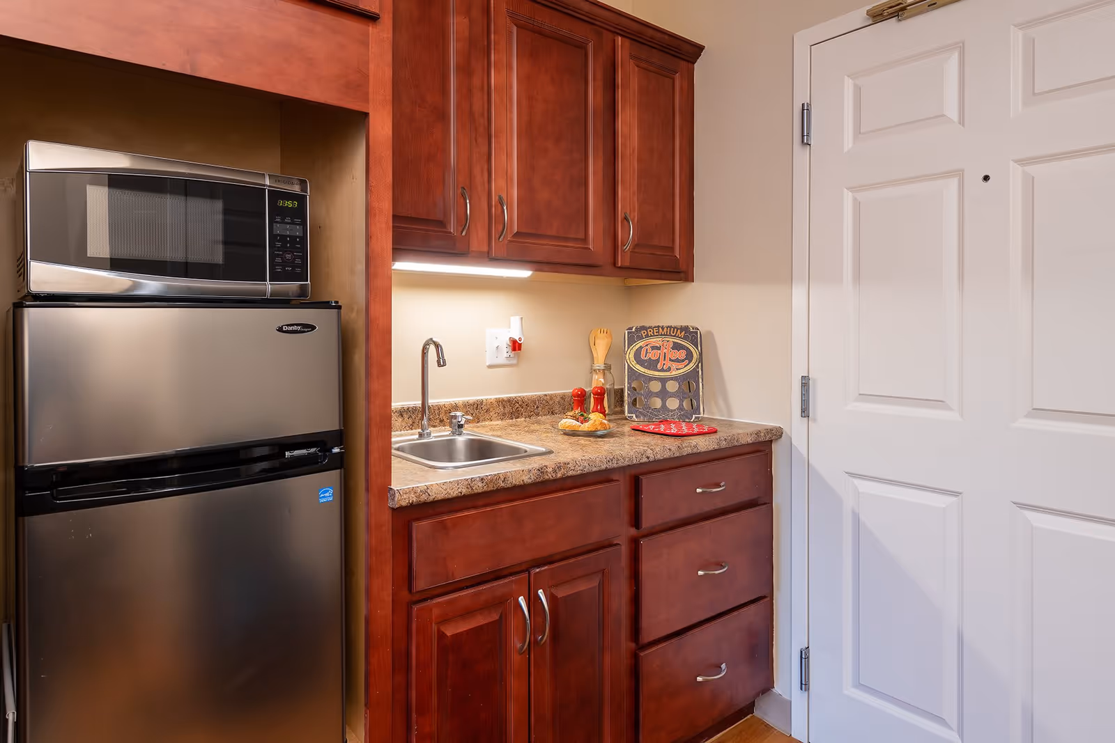 Small kitchenette with a stainless steel refrigerator and microwave, wood cabinets, sink and countertop next to a white door.