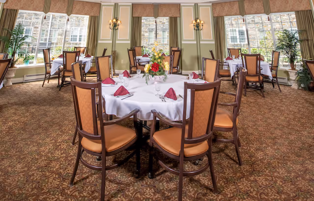 A dining room with round tables covered in white tablecloths, each set with folded burgundy napkins, glassware, and silverware. The room has large windows with beige valances, green walls with white trim, wall sconces, and patterned carpet. There are several wooden chairs with cushioned seats around each table and a floral centerpiece on the nearest table.