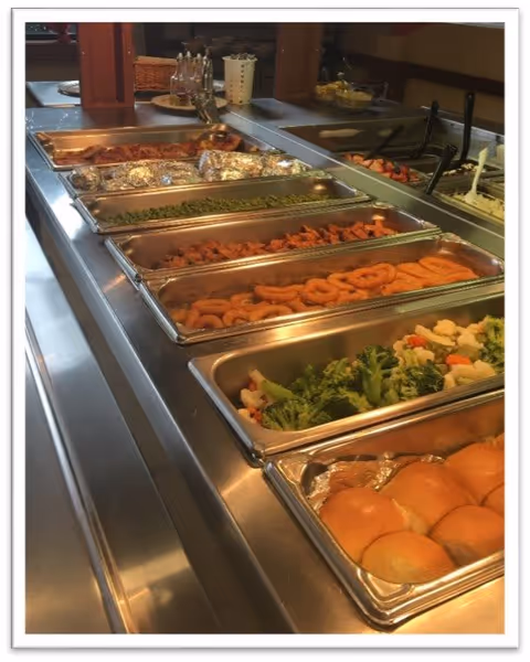 A buffet-style food serving station with trays containing various foods including bread rolls, steamed mixed vegetables, cooked carrots, green beans, and other hot dishes. Serving utensils are visible, and the background shows additional food items and containers.