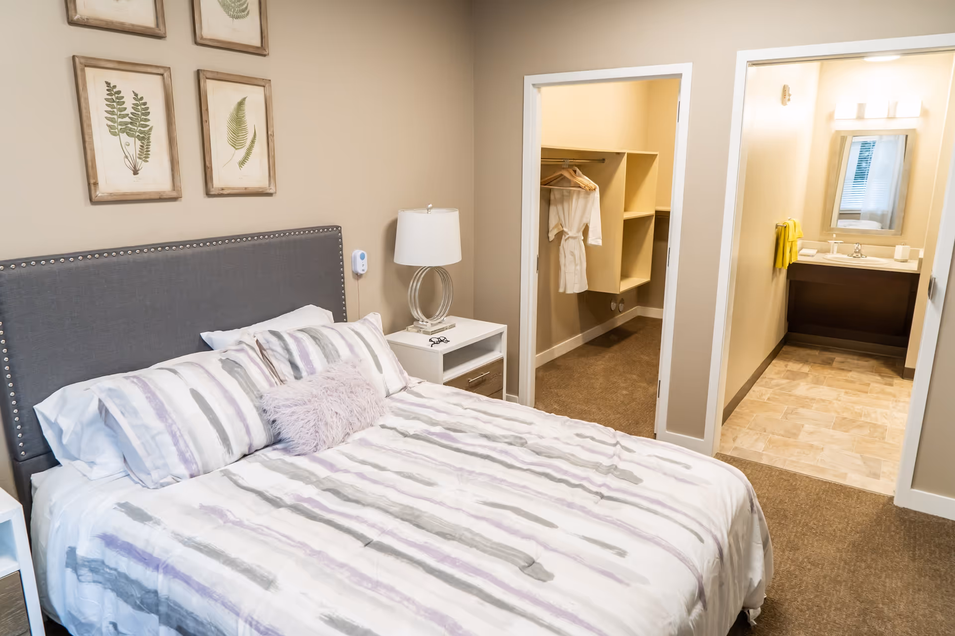 A bedroom with a neatly made bed featuring a striped comforter in shades of gray and purple, a gray upholstered headboard with nailhead trim, and a white nightstand with a modern lamp and eyeglasses. On the wall above the bed are three framed botanical prints. To the right, there are two doorways leading to a walk-in closet with hanging robes and a bathroom with a sink, mirror, and yellow towels.