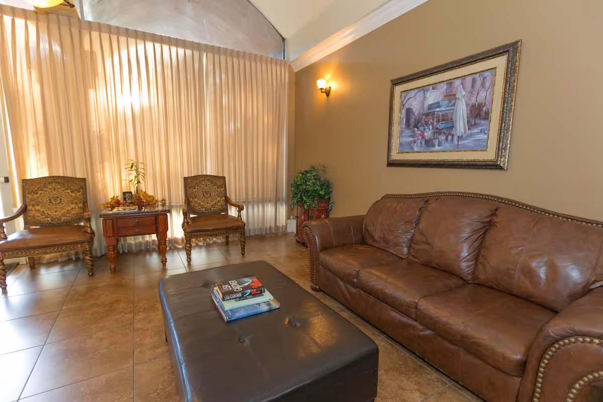 A cozy living room with a brown leather sofa, two ornate wooden chairs with patterned cushions, a small wooden side table with decorative items, a large ottoman with books on top, a potted plant in the corner, a framed painting on the wall, and large windows covered with beige curtains.