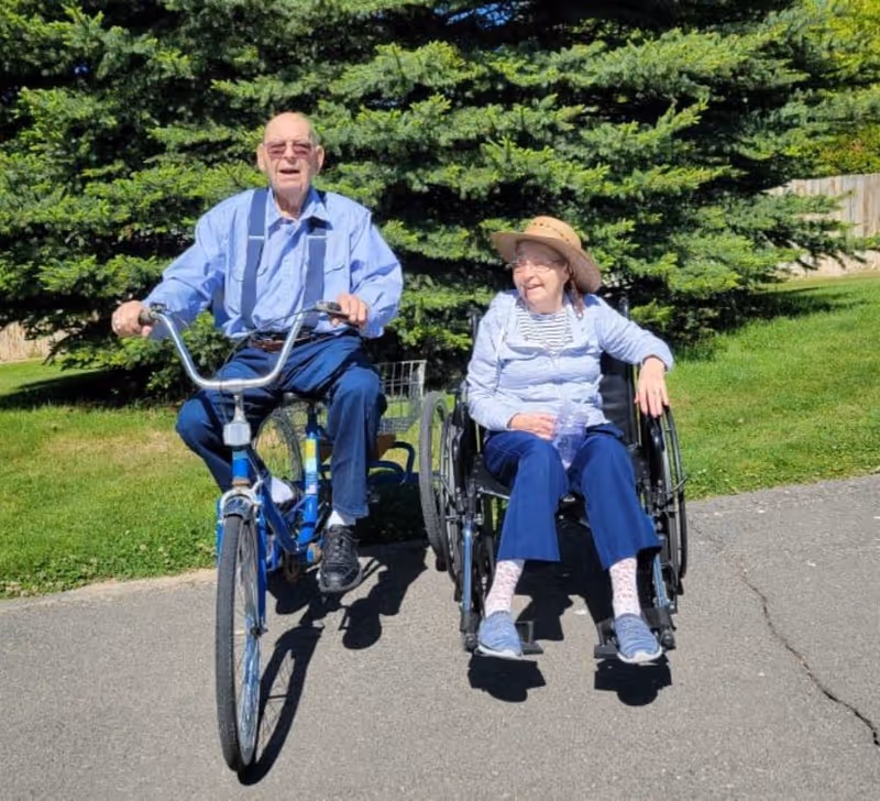 An elderly man riding a blue bicycle next to an elderly woman sitting in a wheelchair on a paved path with green grass and tall evergreen trees in the background. Both are outdoors on a sunny day, and the woman is wearing a wide-brimmed hat and smiling.